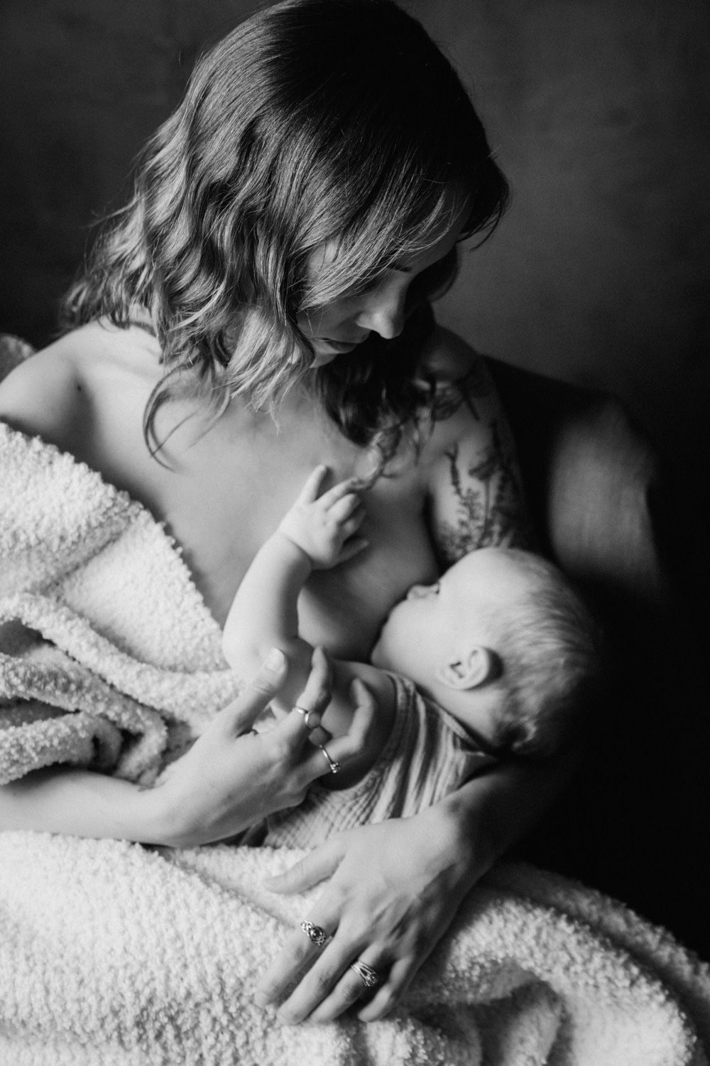 Black and white photo of a tattooed mother breastfeeding her newborn baby wrapped in a soft blanket.