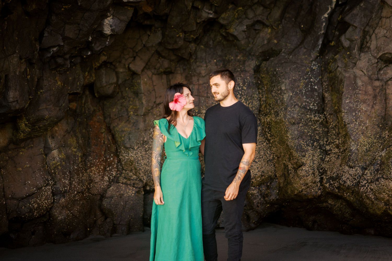 A couple stands romantically before a dark rocky cave wall, woman in green dress with pink hair flower, man in black outfit.