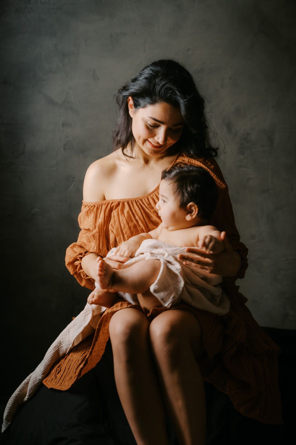 A young mother in an orange dress tenderly holds her newborn baby in a moody studio portrait.