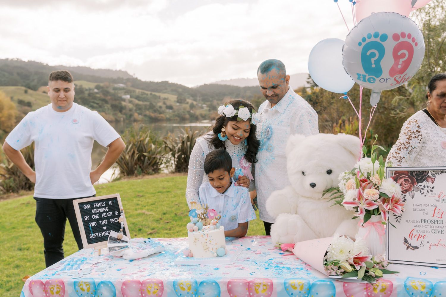 Family gathered outdoors at a gender reveal party with balloons, teddy bear, and decorated table on a green hillside.