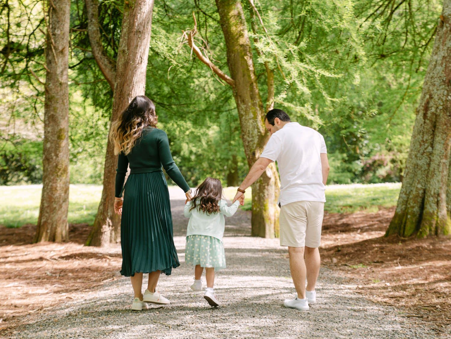 A family of three walks hand-in-hand down a tree-lined path in a lush green park.