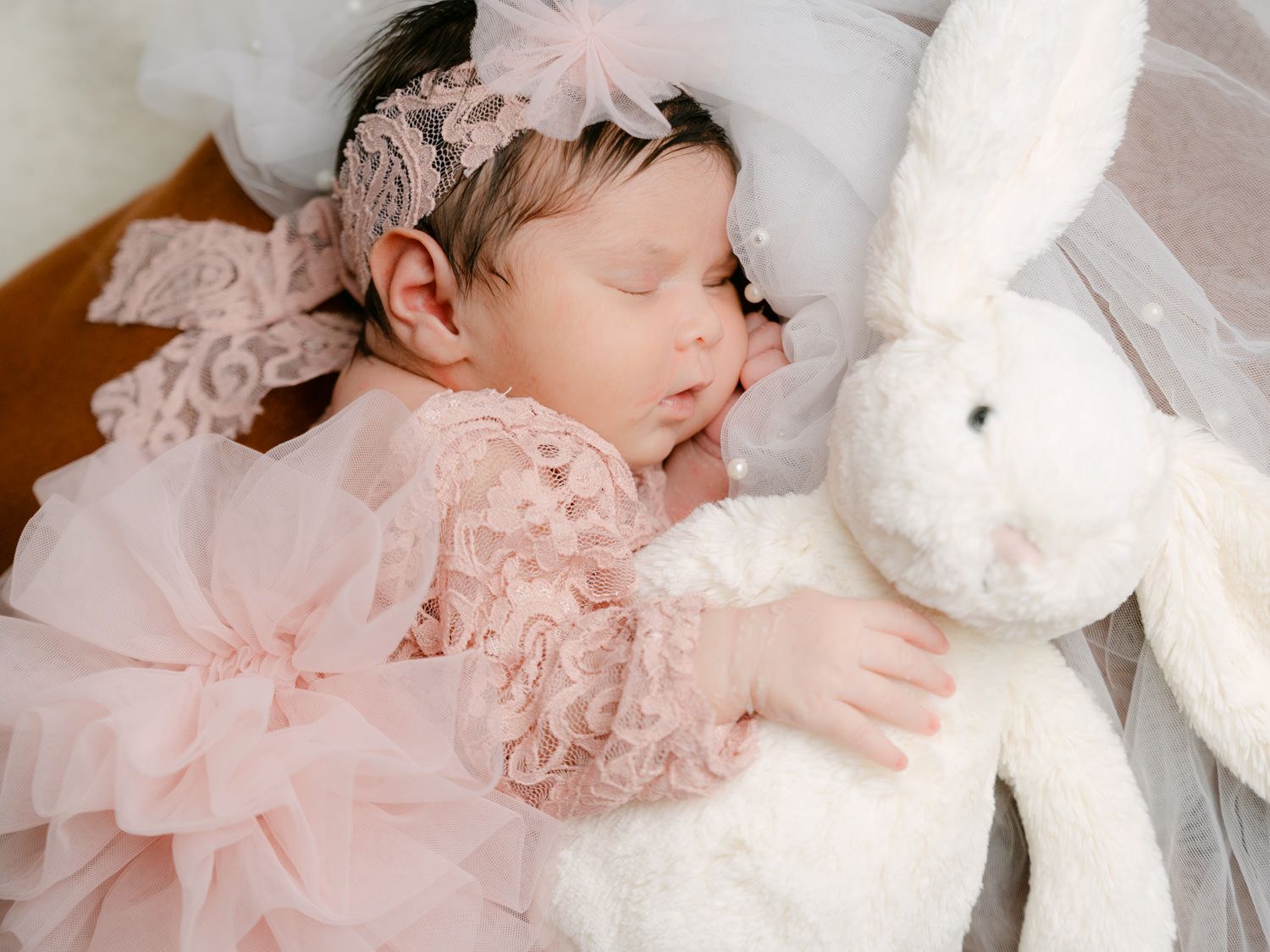 Sleeping newborn baby girl in pink lace dress with floral headband cuddling white bunny stuffed animal.
