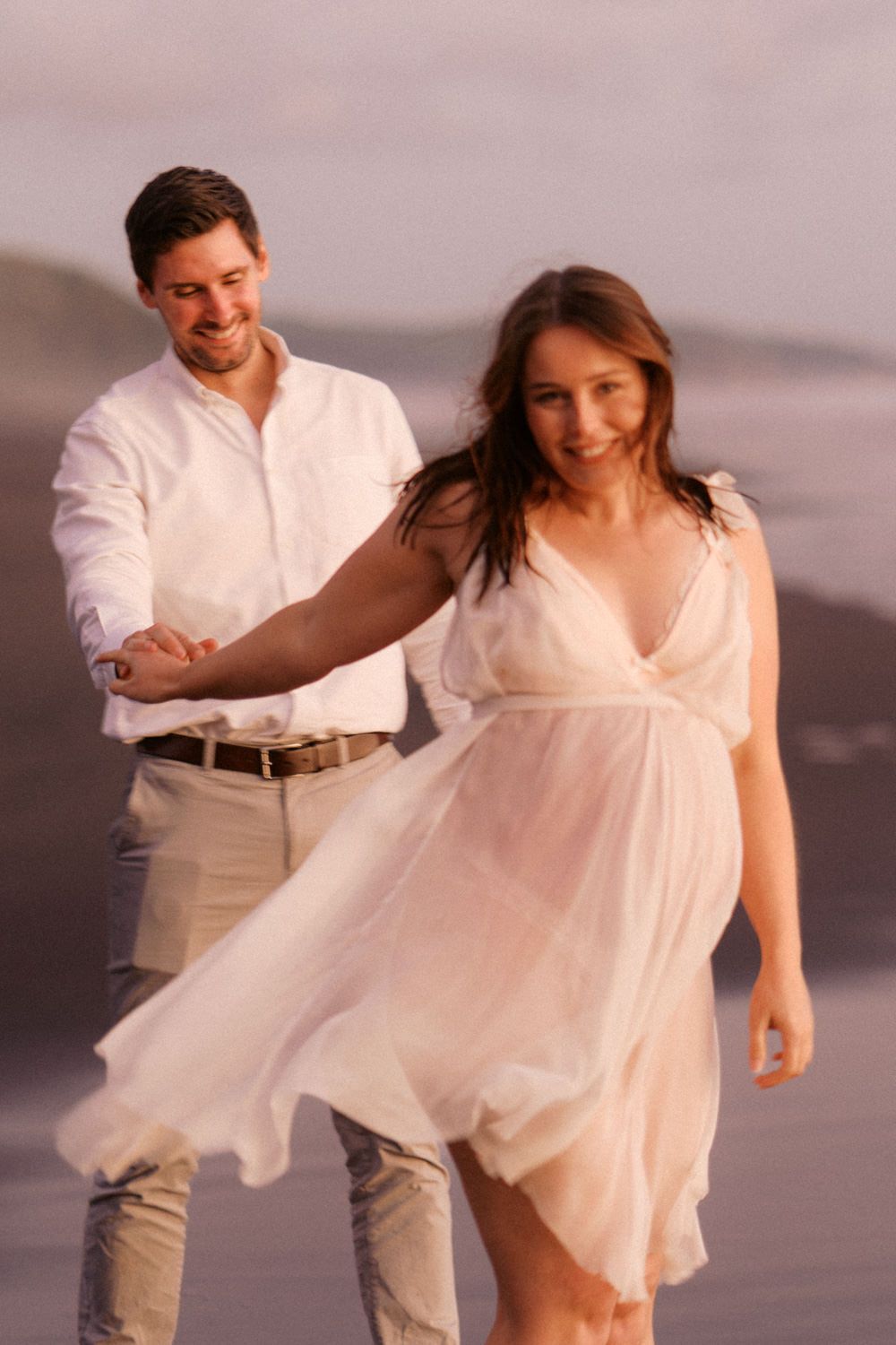 Couple holding hands on a beach at sunset, woman in flowing white dress, man in white shirt smiling.