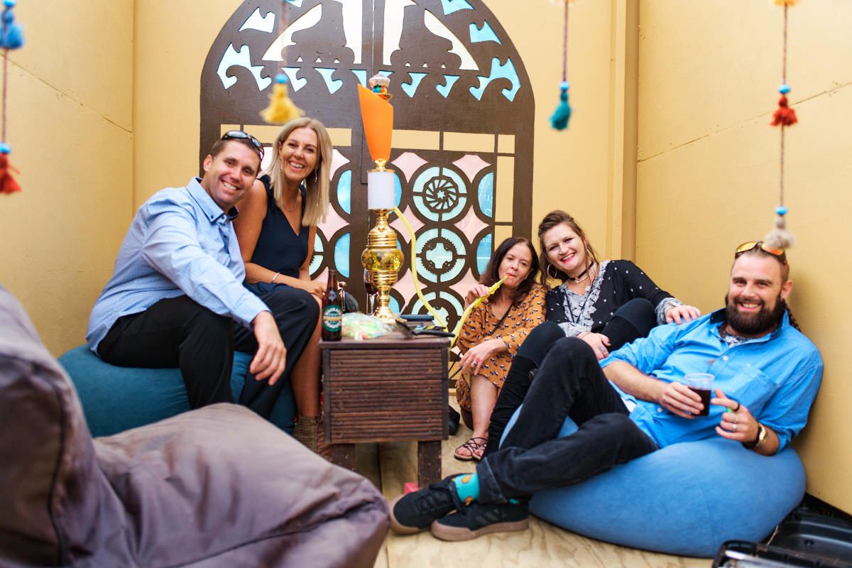 Five friends smiling and relaxing together in a cozy Moroccan-themed lounge with bean bags and decorative arch.