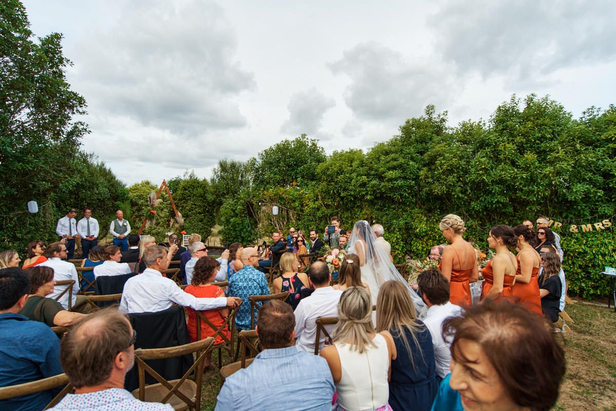 Outdoor wedding ceremony in a lush garden setting with guests seated facing the couple under cloudy skies.