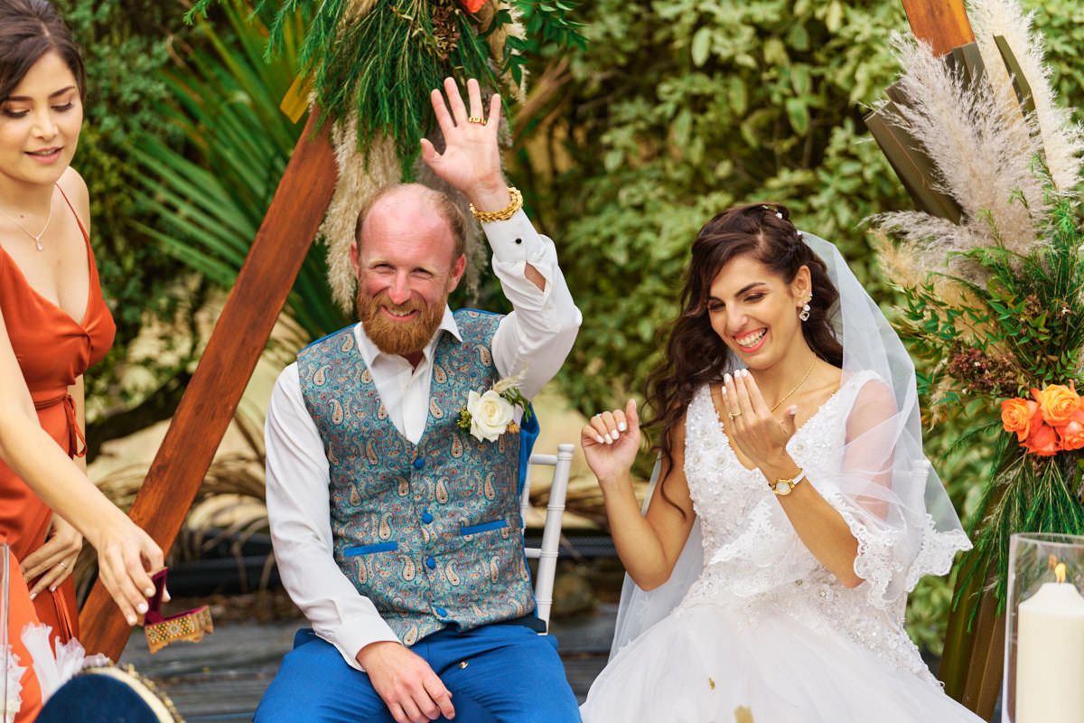 Joyful groom with red beard waves during outdoor wedding ceremony beside smiling bride in white dress.