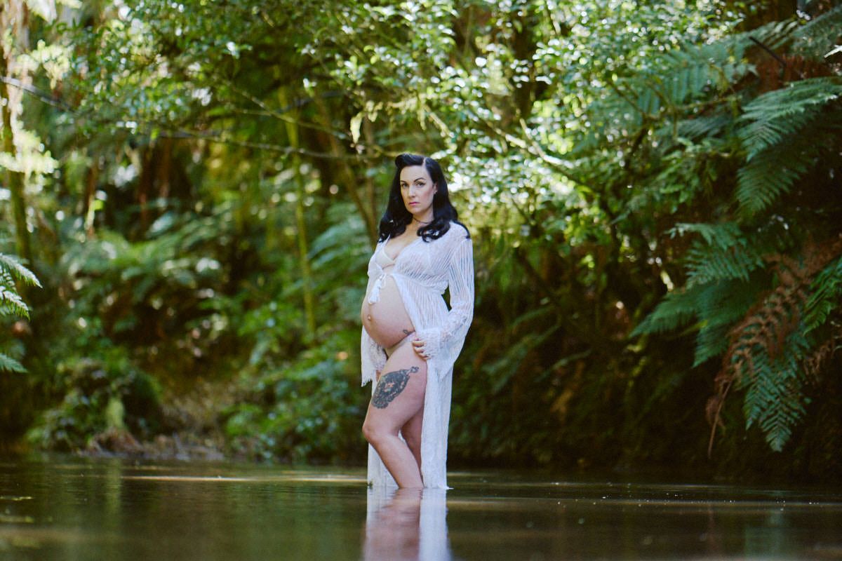 Pregnant woman in white sheer dress standing in a serene forest stream surrounded by lush green ferns and trees.