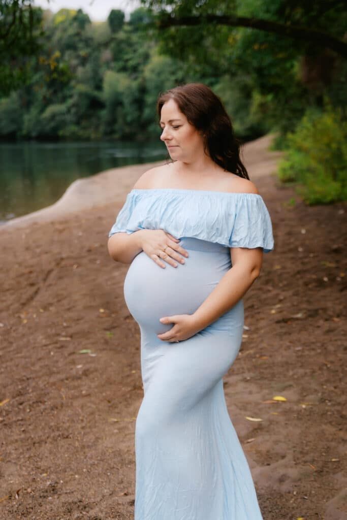 Pregnant woman in light blue off-shoulder gown cradling her baby bump by a serene riverside.