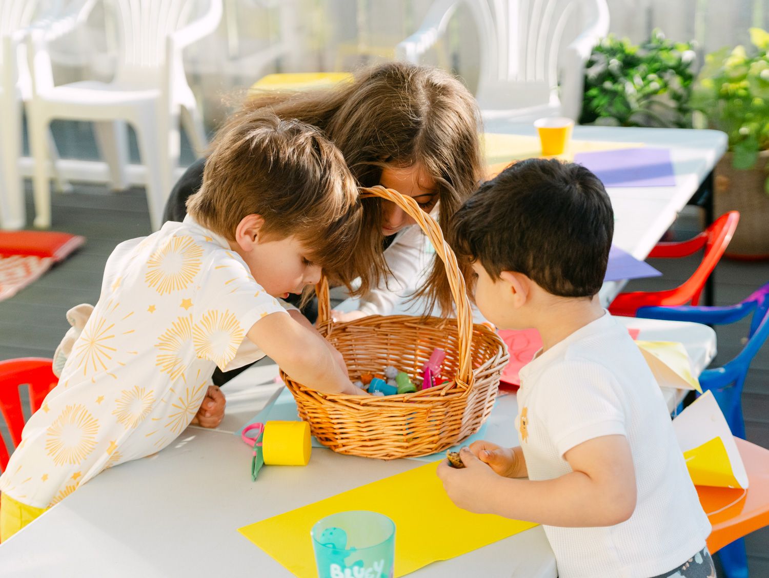 Three young children gathered around a wicker basket on a white table at a colorful outdoor party.