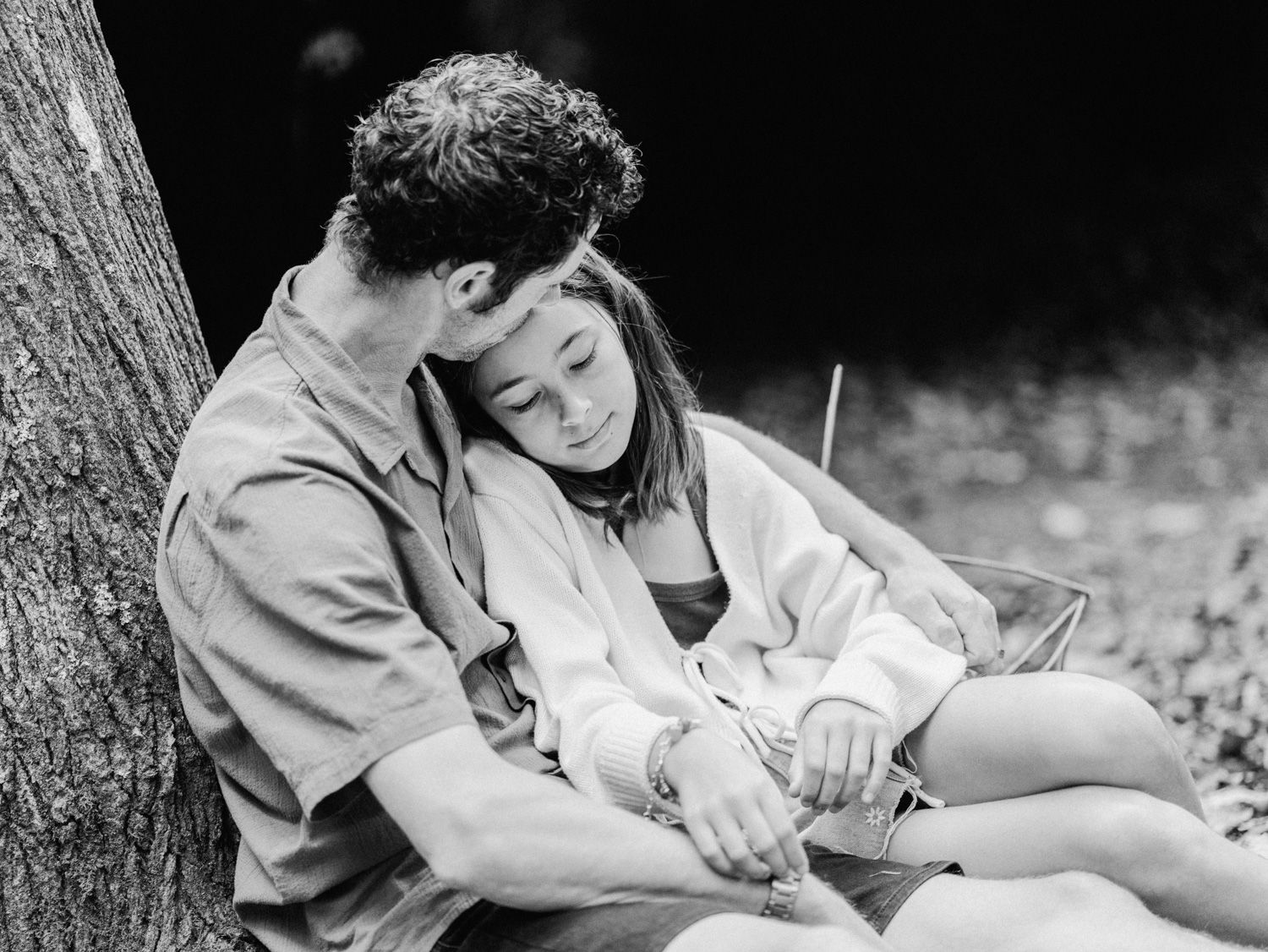 Black and white photo of a couple sitting against a tree, woman resting her head on mans shoulder outdoors.