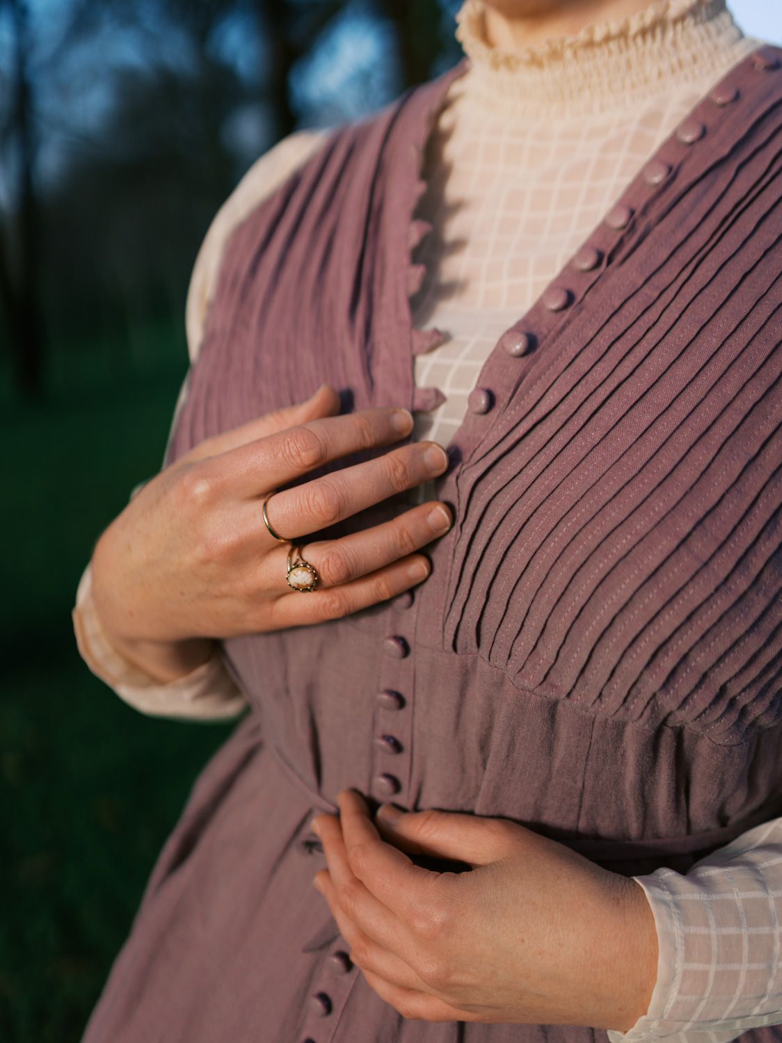 Close-up of a woman wearing a mauve pleated vest with buttons over a cream lace blouse, adorned with a gold ring.