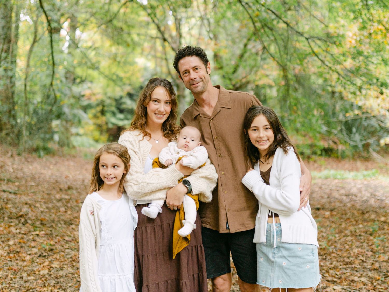 Happy family of five posing outdoors in autumn forest, parents with three children including a baby, smiling among fall foliage.