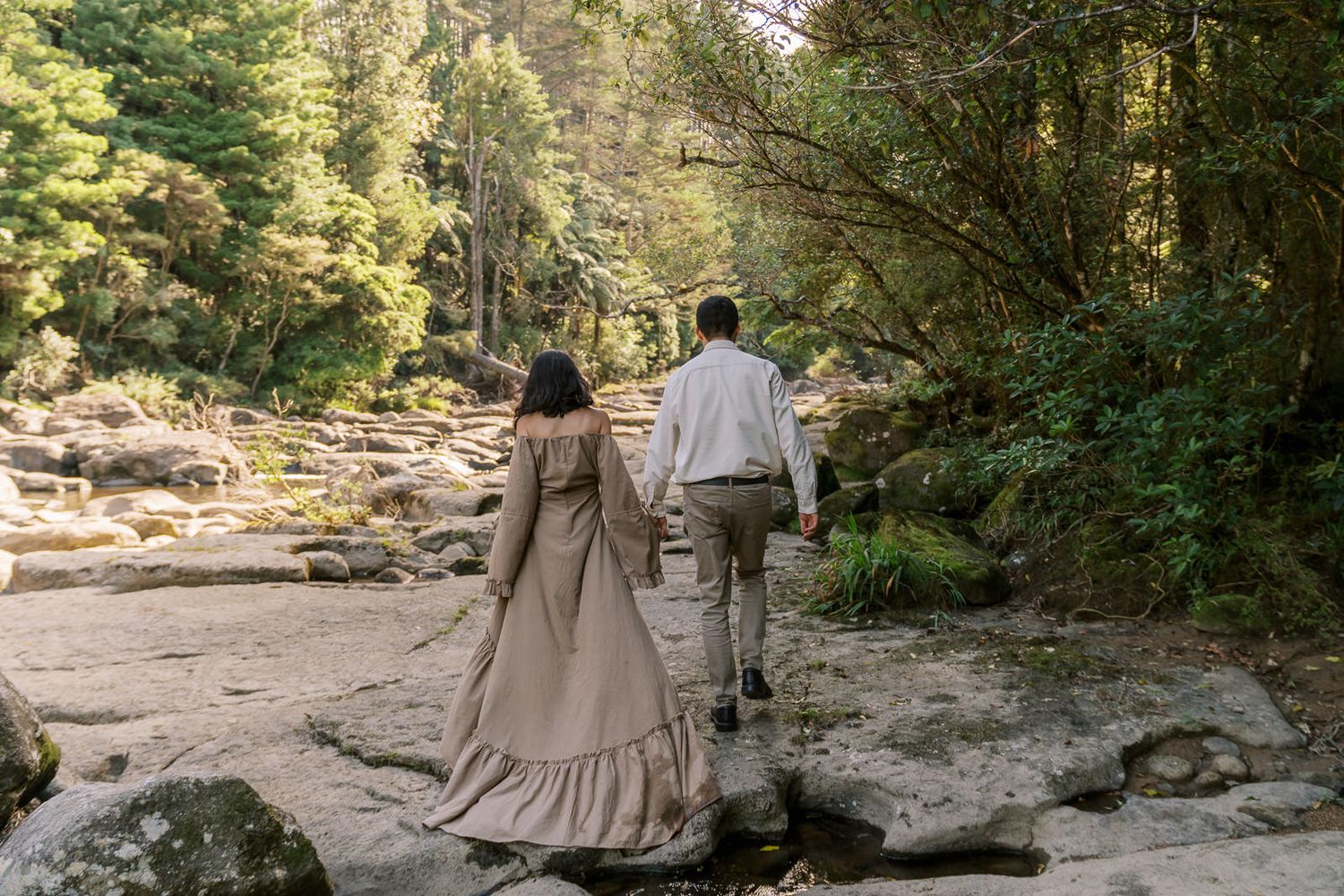 Couple holding hands walking on rocky riverbed surrounded by lush forest trees in nature.