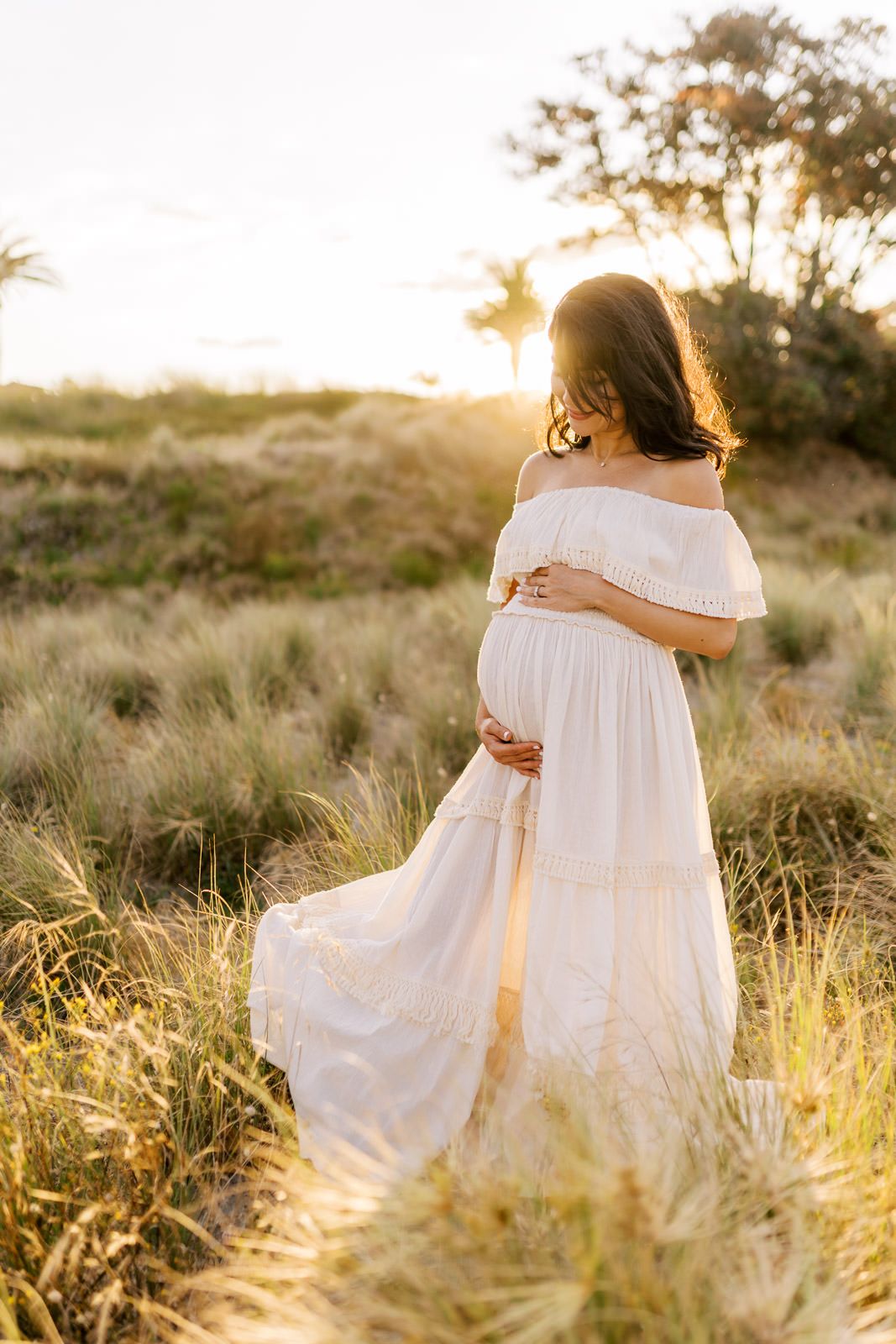 Pregnant woman in white off-shoulder dress cradling belly in golden field at sunset.