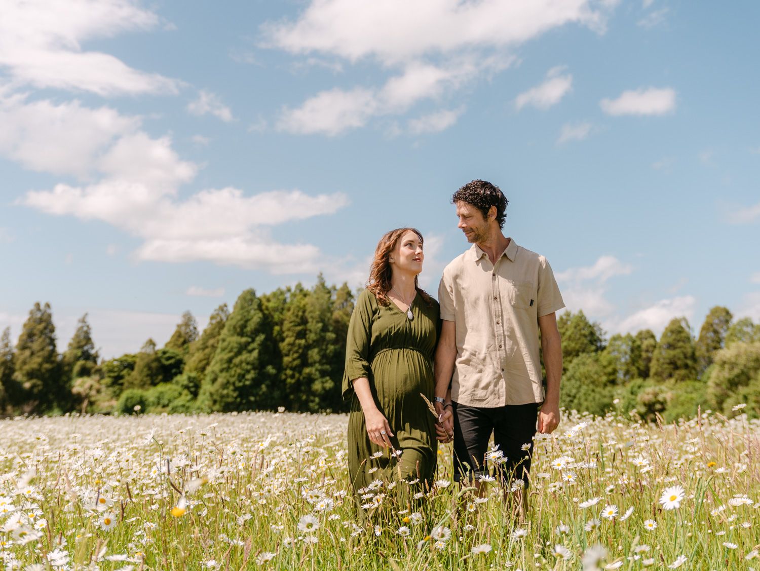 Pregnant couple holding hands in a wildflower meadow with blue sky and trees in the background.