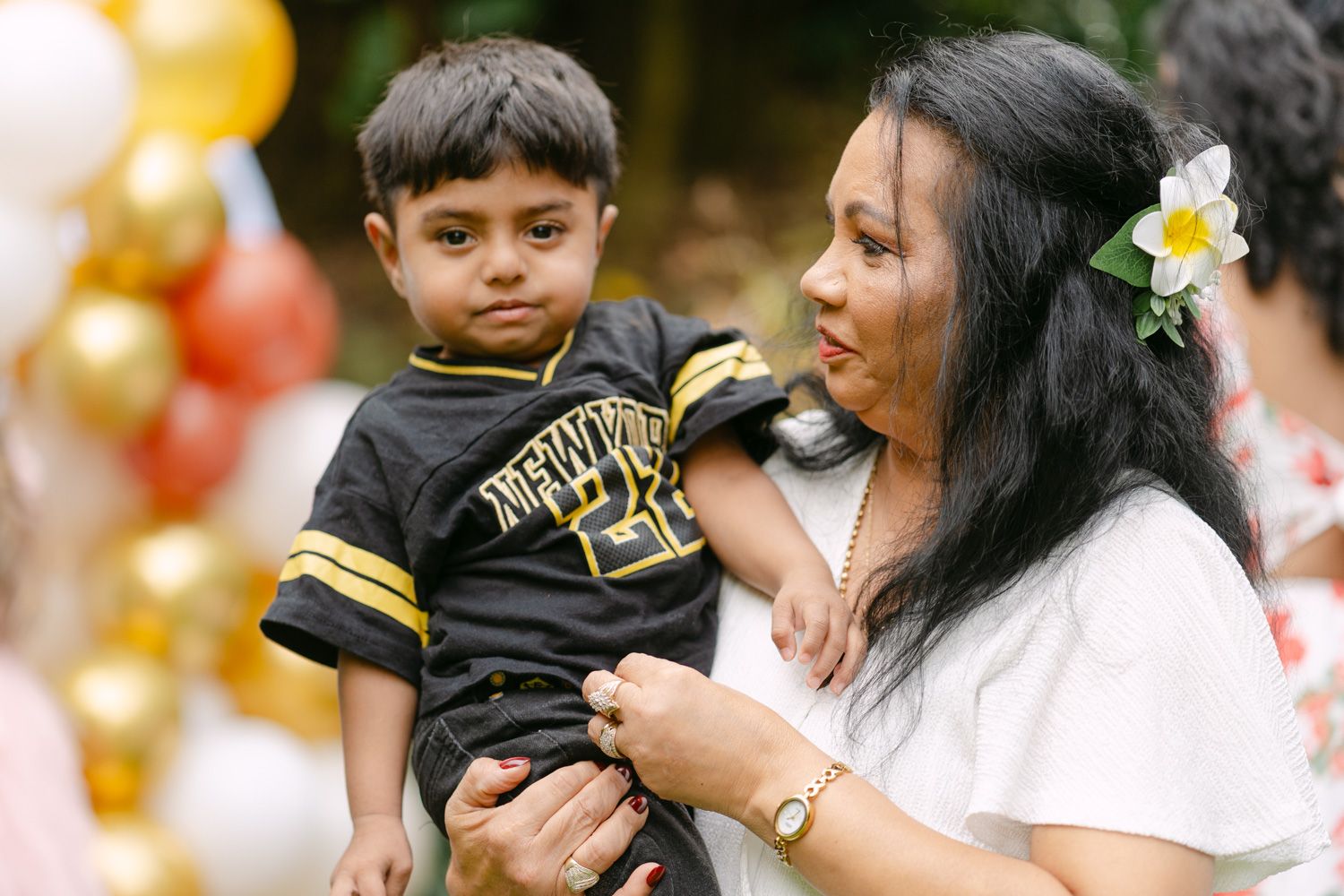 Woman holding a young boy at a festive outdoor party with red, white, and gold balloons in the background.
