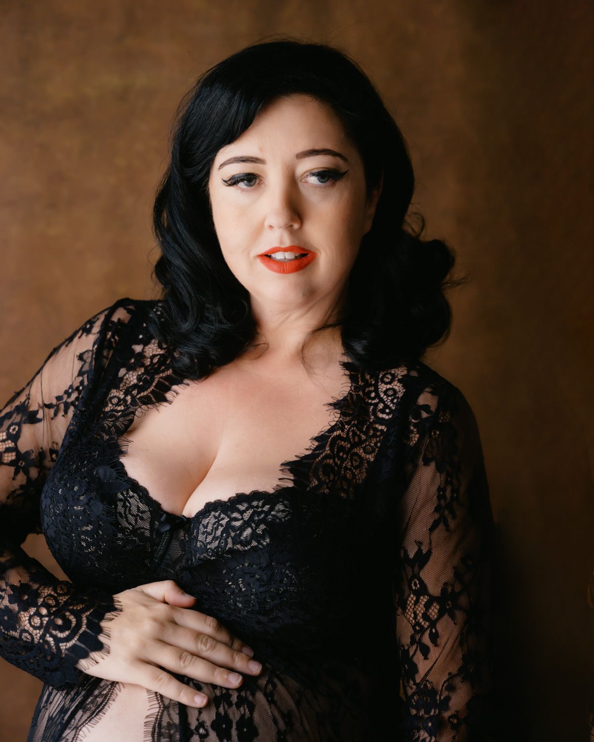 Woman in black lace lingerie robe posing confidently against a warm brown studio backdrop.