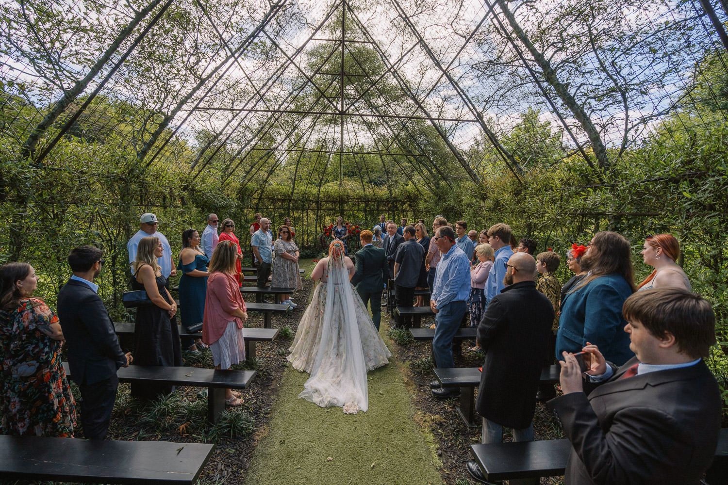 Bride walks down the aisle in a stunning greenhouse wedding venue surrounded by lush greenery and seated guests.