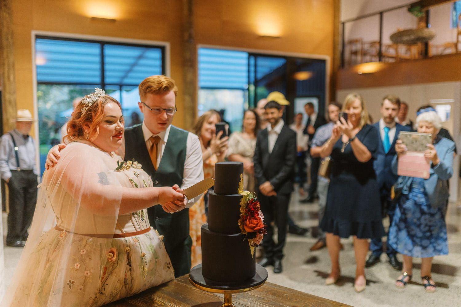 Bride and groom cutting a black wedding cake surrounded by guests at a rustic wedding reception venue.