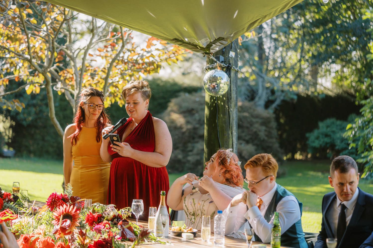 Two women give a toast at an outdoor wedding reception table adorned with red flowers and champagne glasses.