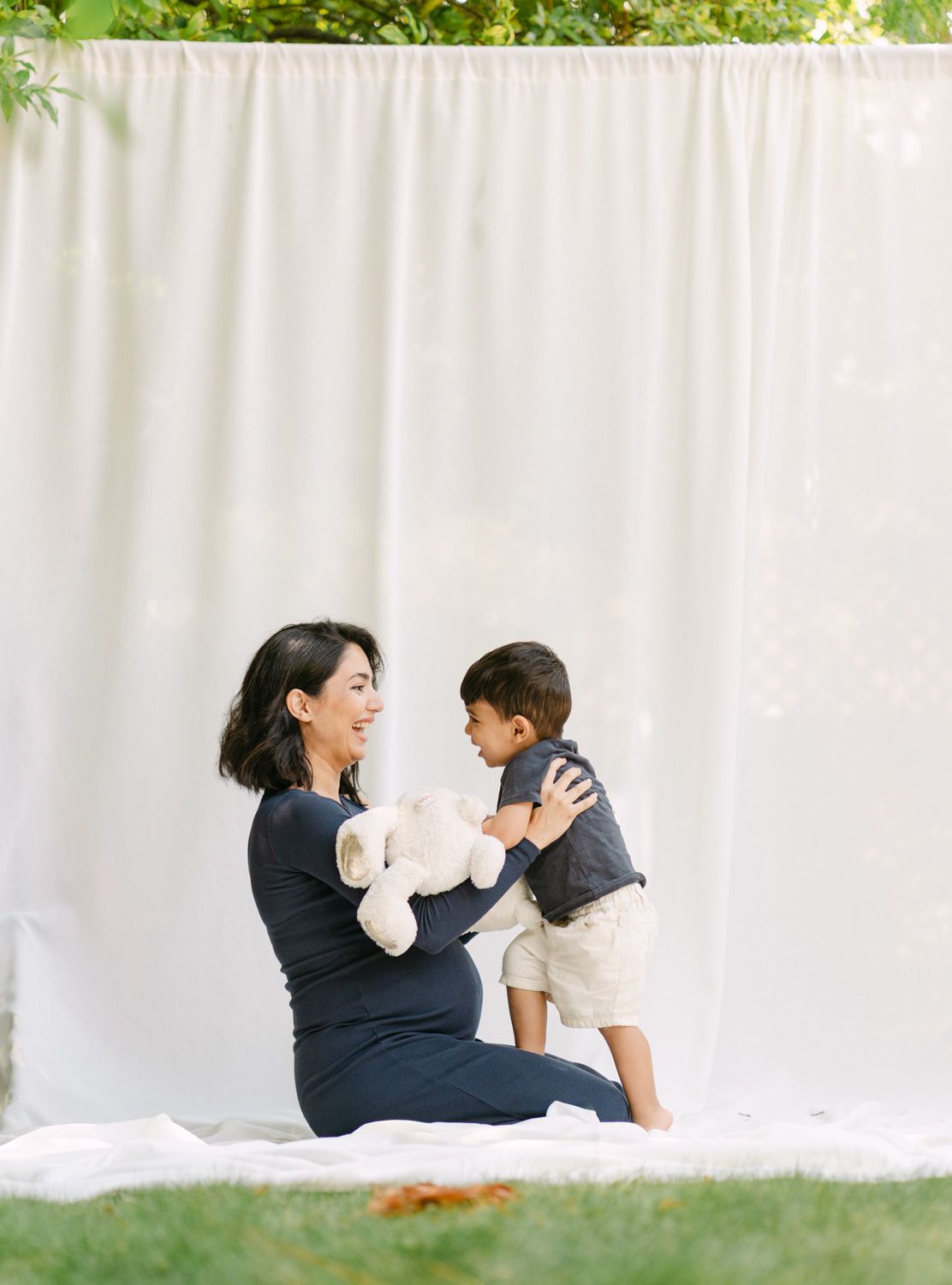 Pregnant woman sitting on white blanket outdoors, smiling at toddler boy holding a white teddy bear against draped backdrop.