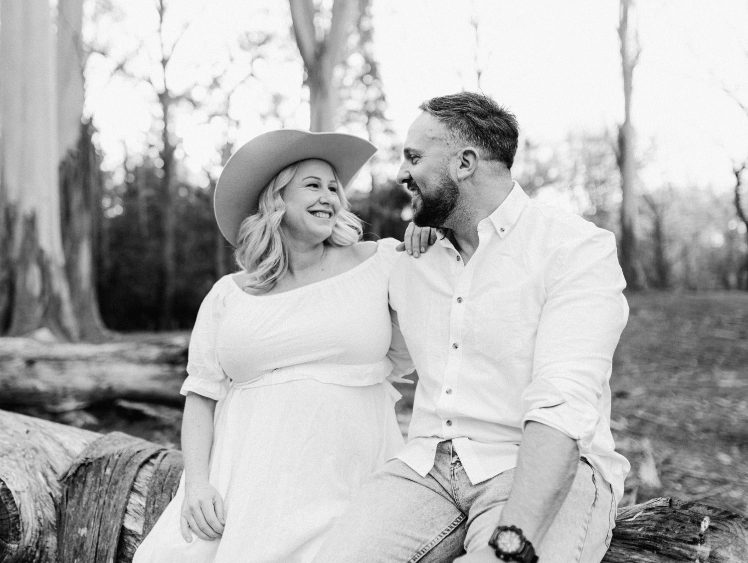 A smiling couple sits on a log in a wooded area, the woman wearing a white dress and cowboy hat, the man in a white shirt.