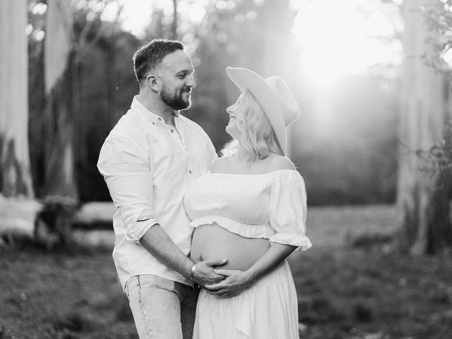 Black and white maternity photo of couple in white outfits embracing outdoors with sunlit trees in background.