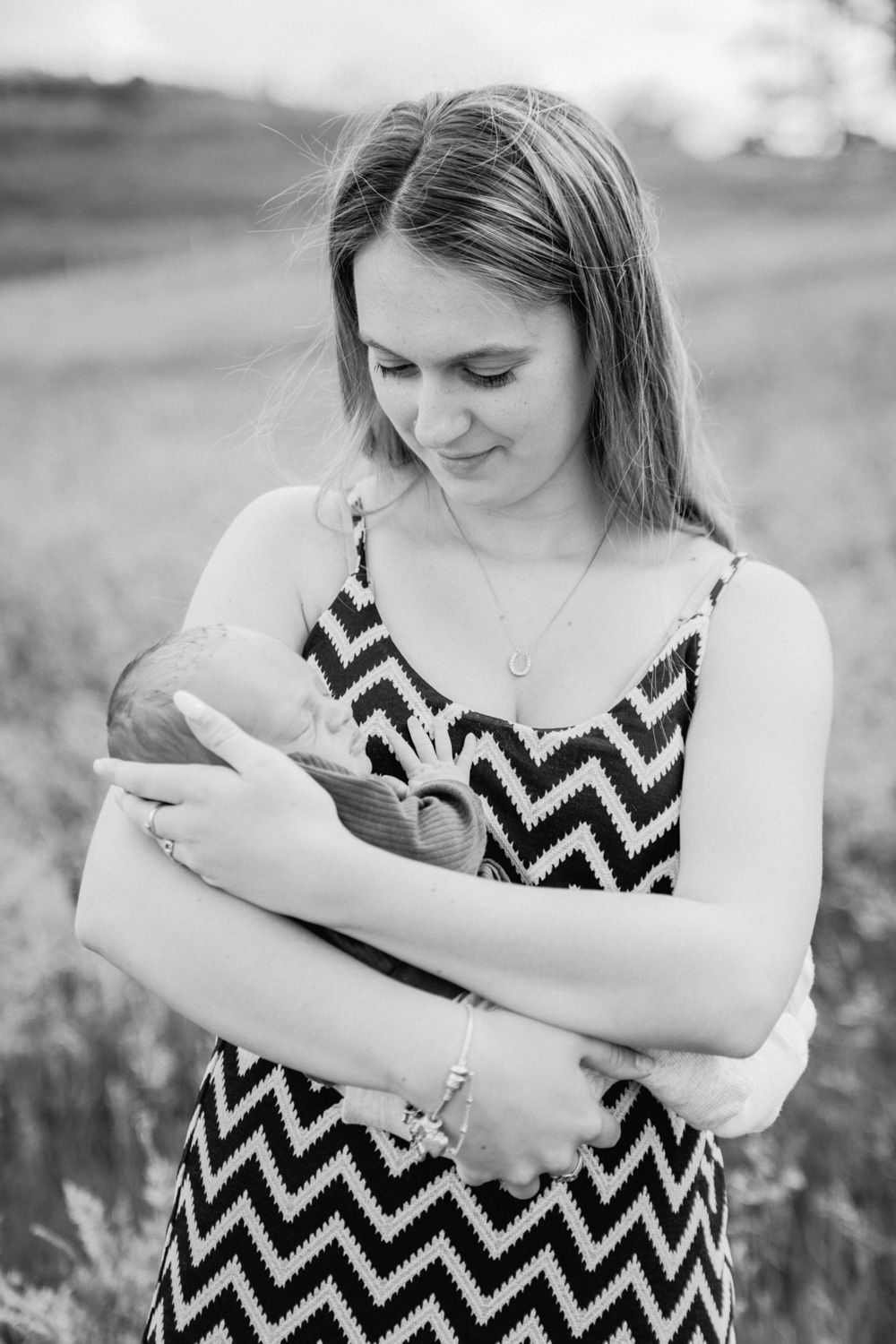 Black and white photo of a young mother in chevron dress tenderly cradling her newborn baby outdoors in a field.