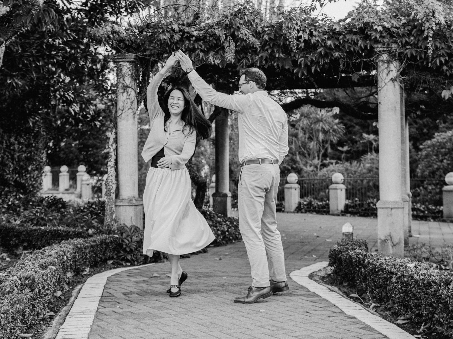 Couple dancing together on a brick garden path surrounded by lush greenery and stone columns in black and white.