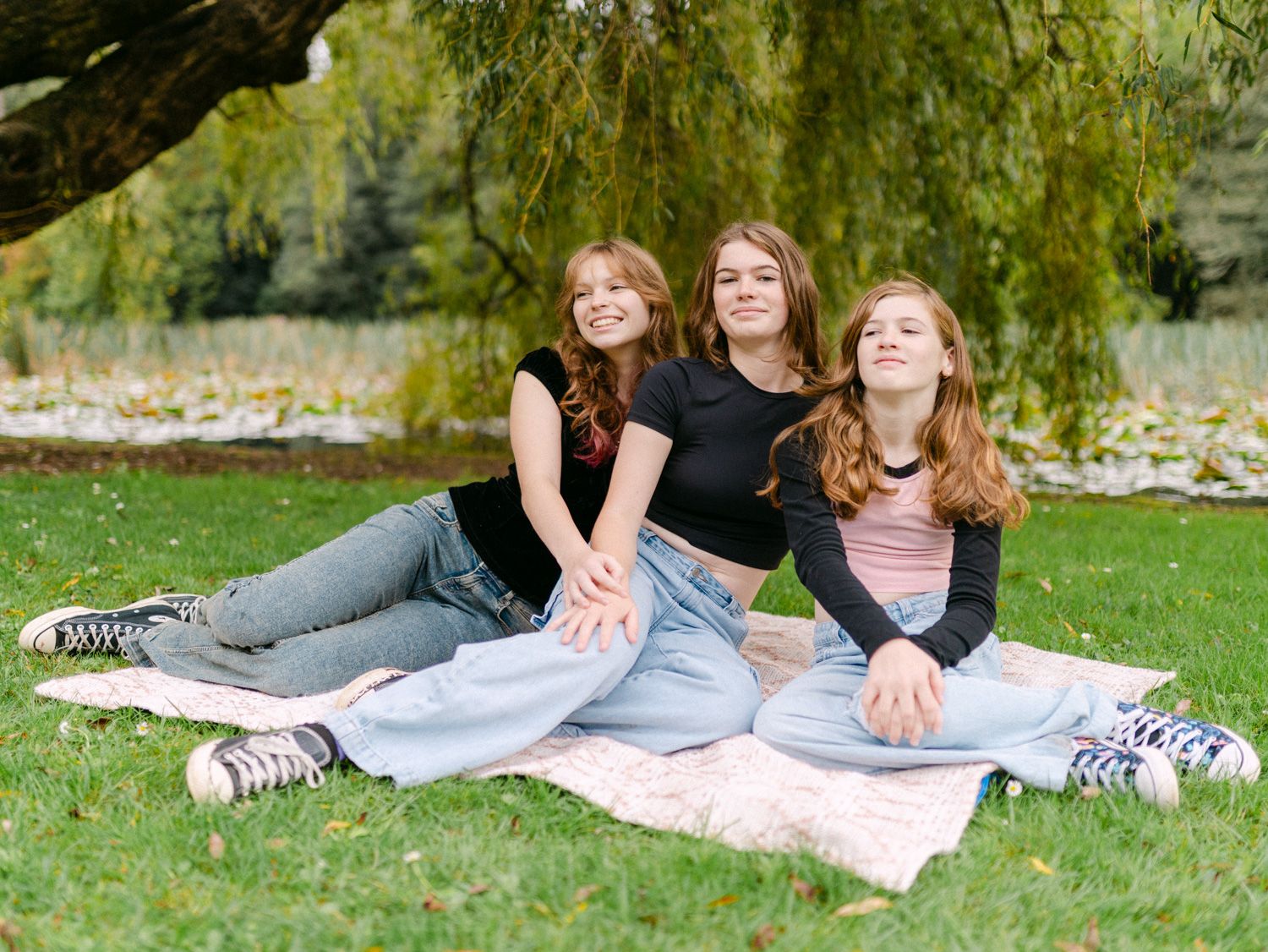 Three teenage girls sitting together on a blanket under a willow tree in a park, smiling and posing for a photo.