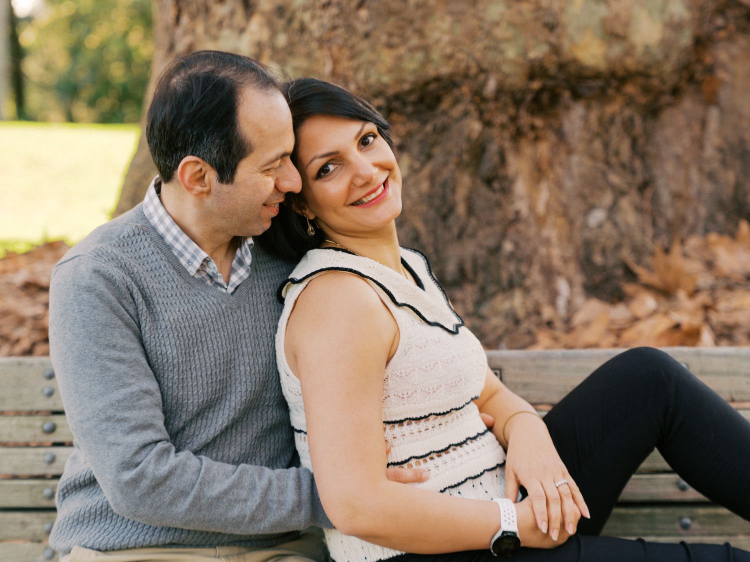 A couple sits on a park bench, the pregnant woman smiling warmly as her partner leans in affectionately.