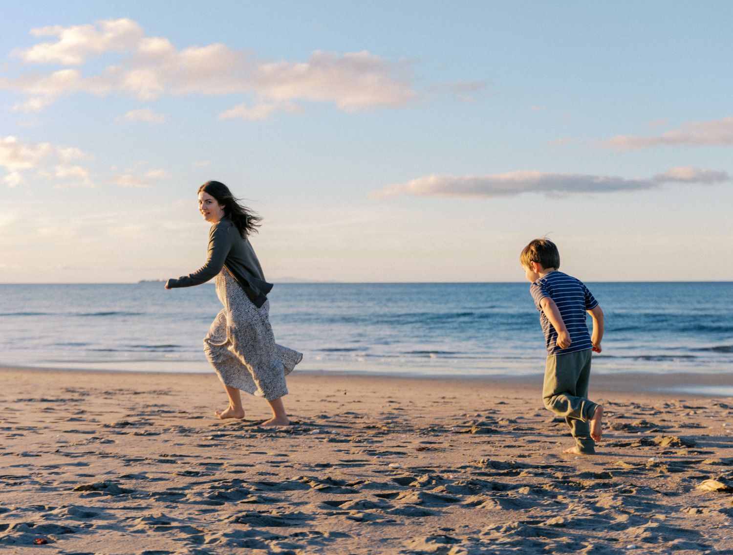 A woman and child playing joyfully on a sunny beach with calm ocean waves in the background.