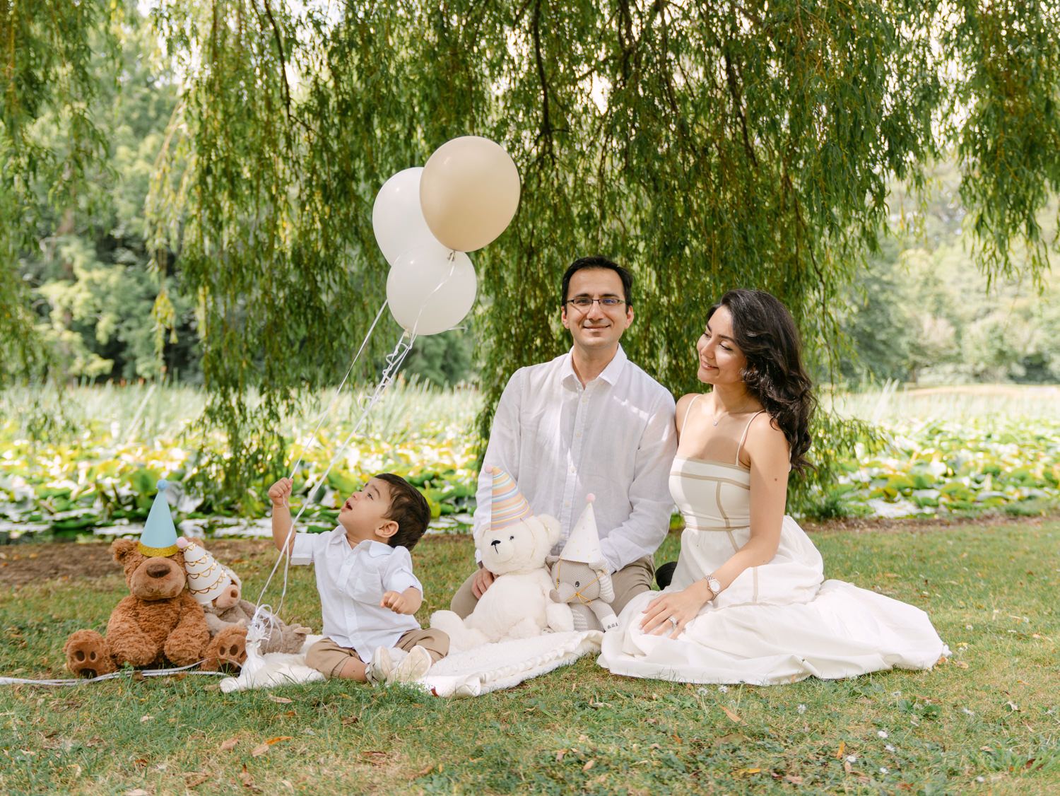 Family in white outfits sitting on grass with teddy bears and balloons during an outdoor park photoshoot.
