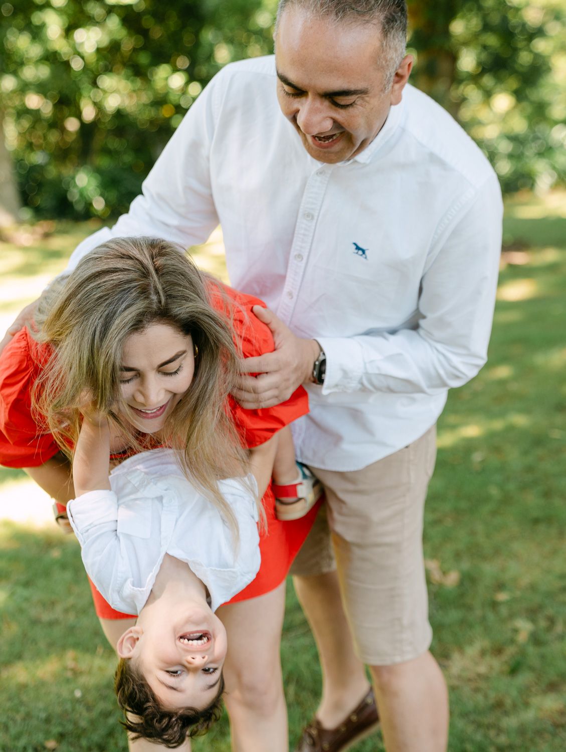 A joyful family plays outdoors, with parents swinging their laughing child upside down in a sunny green park.