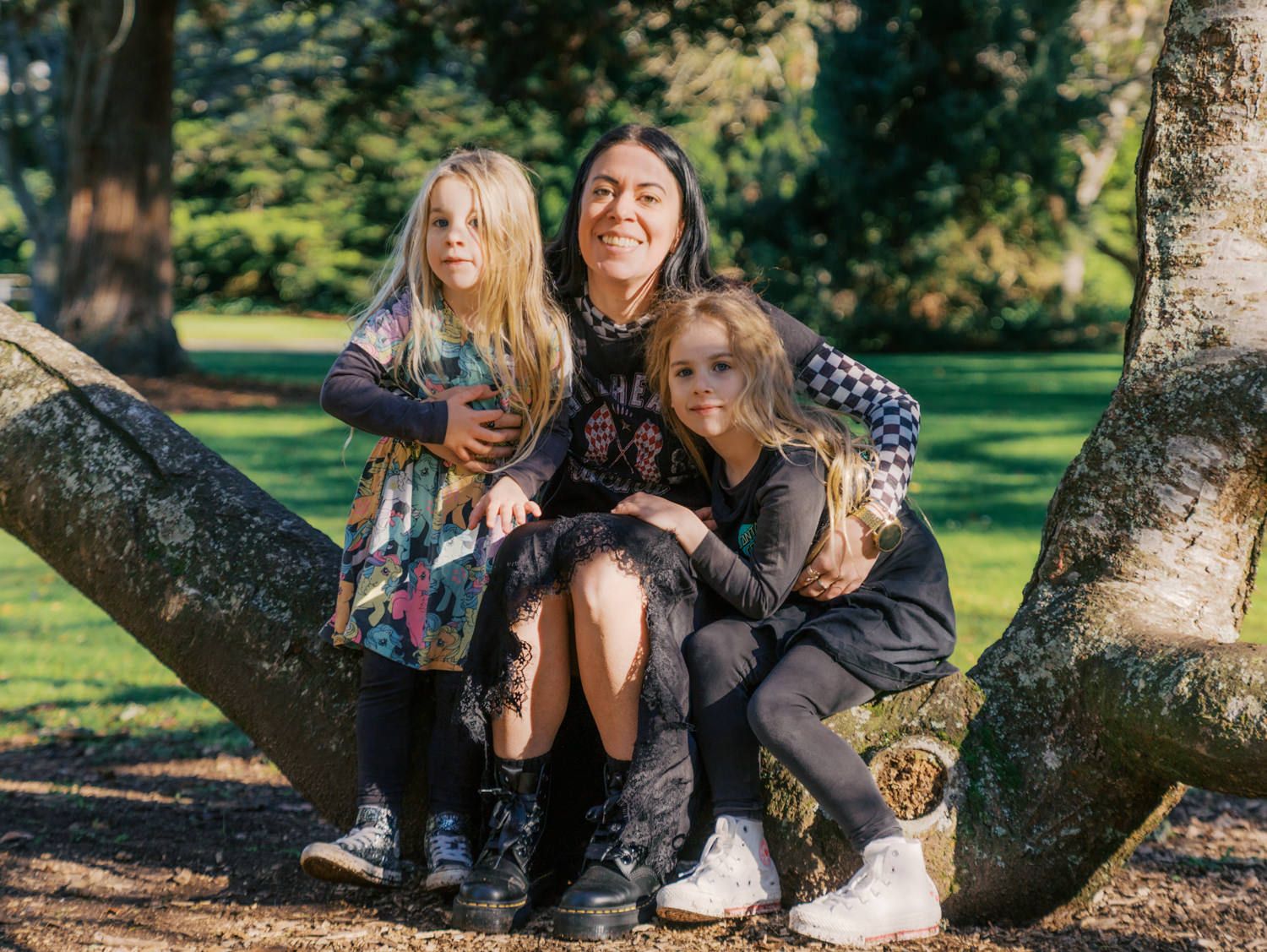 A woman and two young girls pose together on a large tree branch in a sunny park.