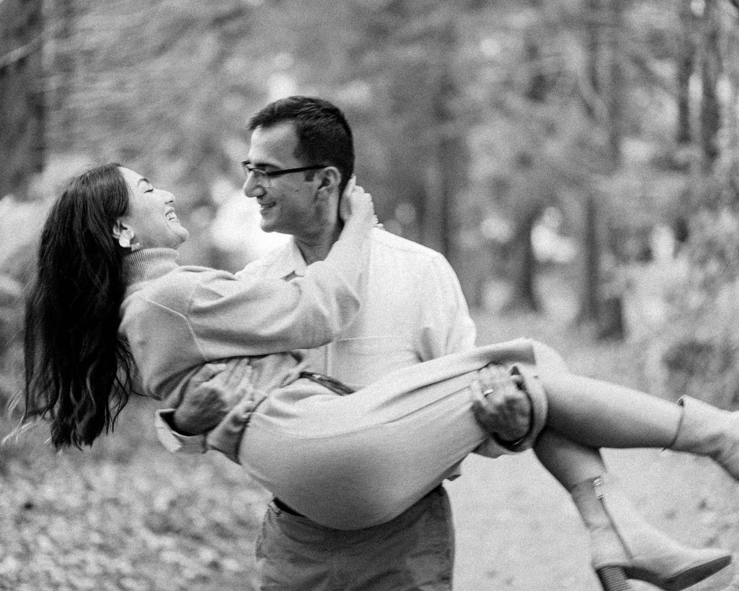 Black and white photo of a man carrying a laughing woman in his arms outdoors among autumn trees.