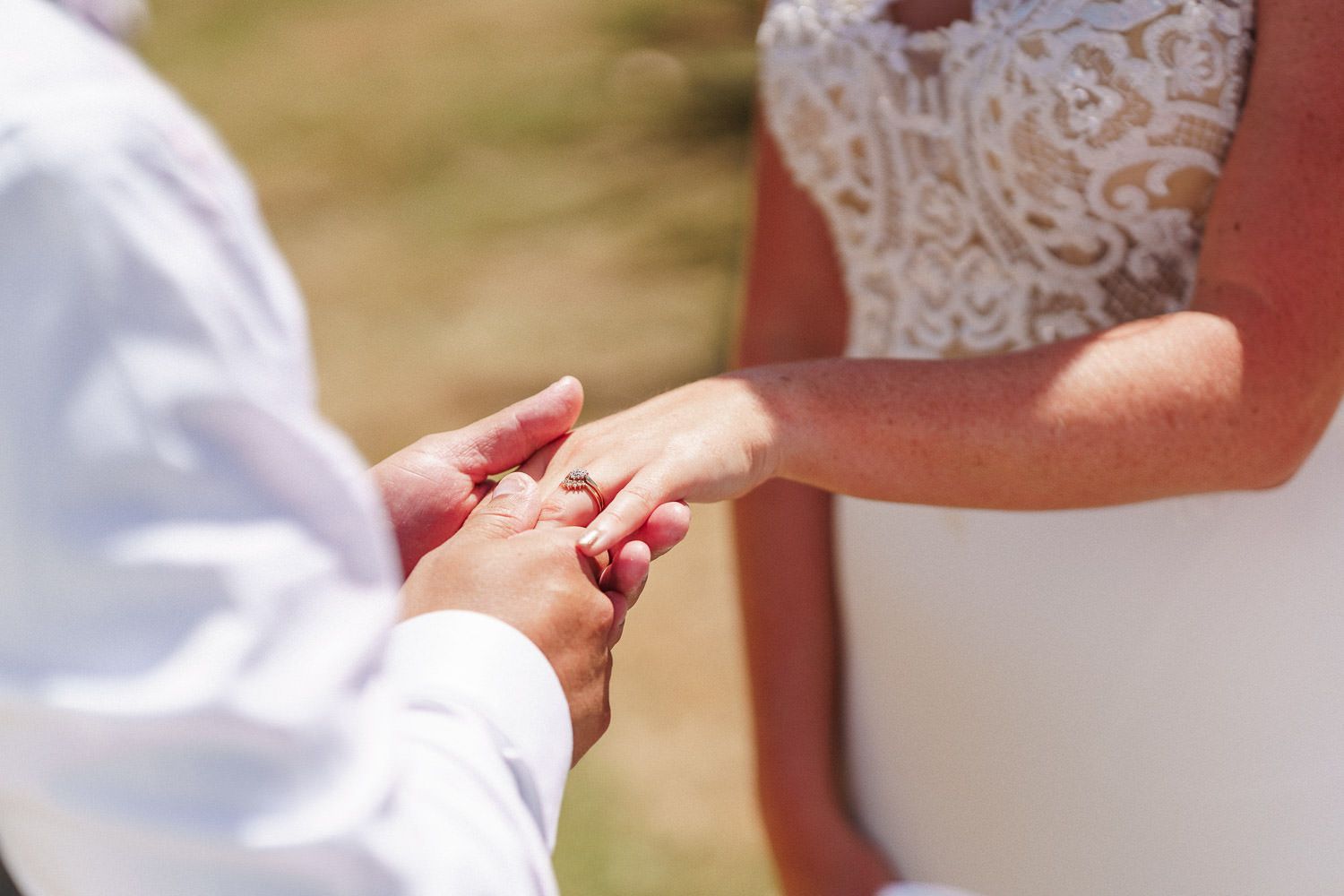 Groom placing a wedding ring on bride's finger during outdoor ceremony, close-up of hands in sunlight.