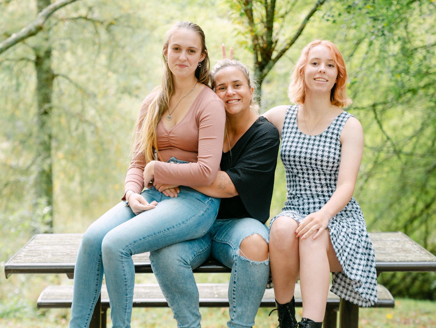 Three young women sitting together on a bench in a lush green park, smiling and posing for a photo.