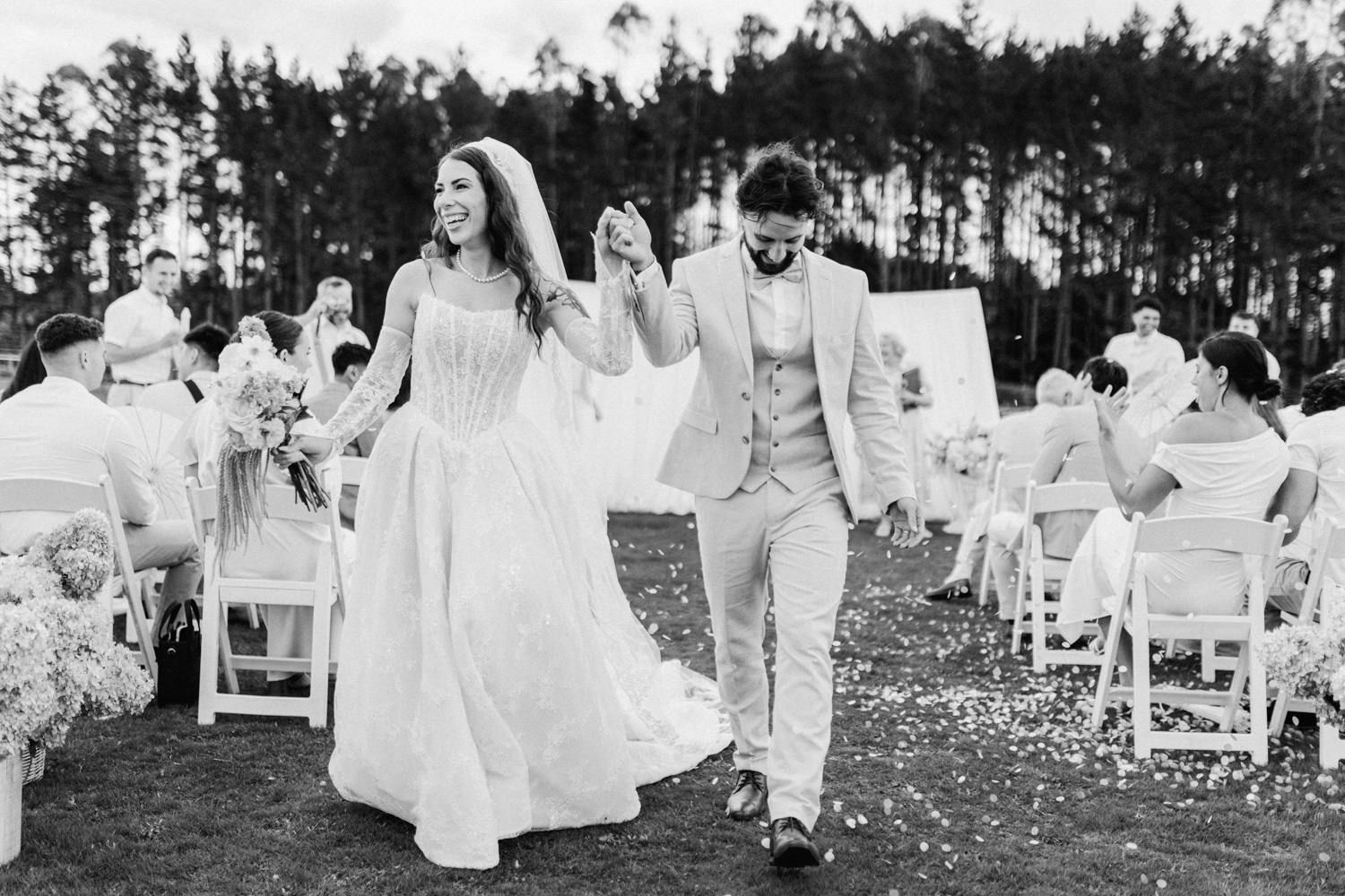 Bride and groom walk joyfully down outdoor aisle after wedding ceremony with guests seated among pine trees.