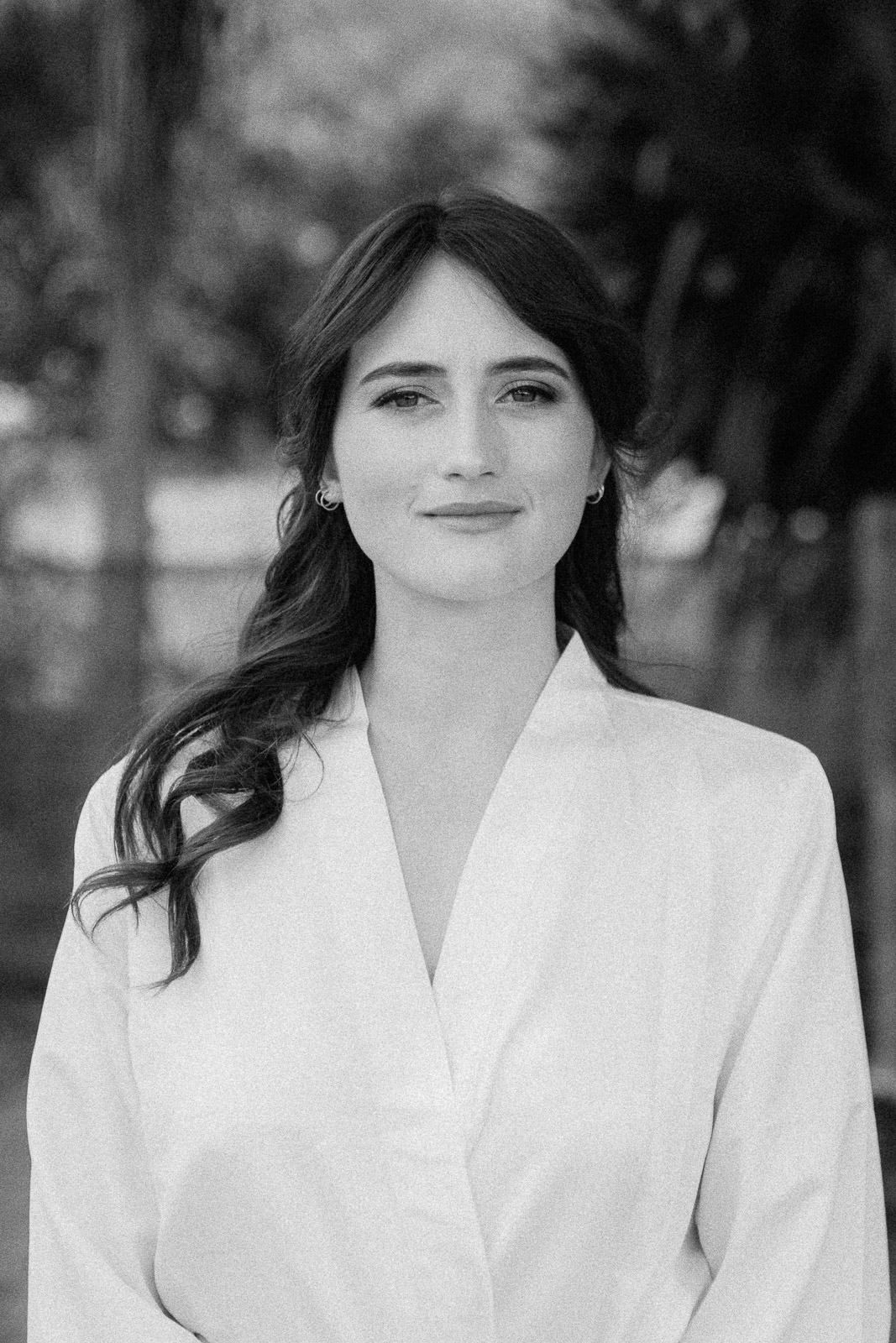 Black and white portrait of a young woman with long dark hair wearing a white blouse, smiling outdoors.