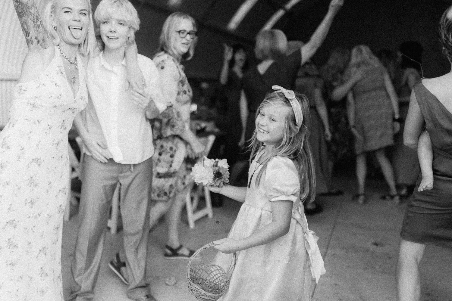 Black and white photo of guests dancing at a wedding reception with a young flower girl holding a basket.