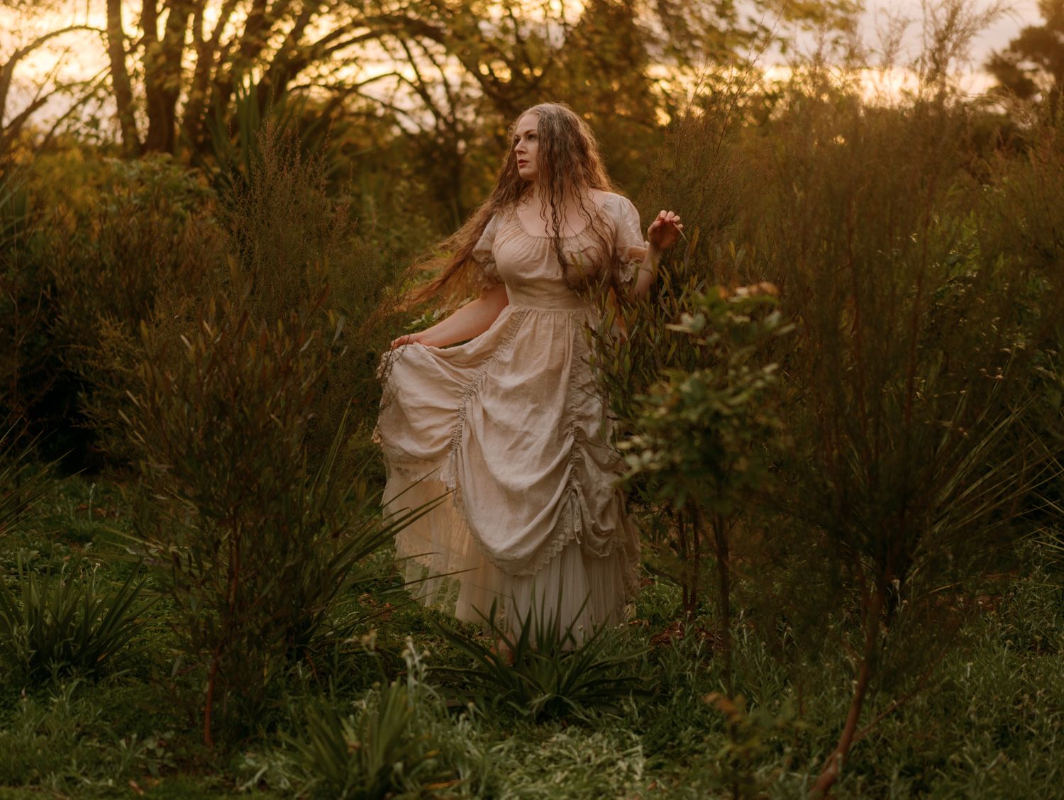 Woman in flowing cream vintage gown standing among lush green bushes in golden sunset light.