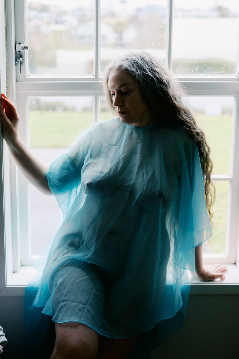 Woman with long hair in sheer blue wrap leans against white window frame, looking down pensively.