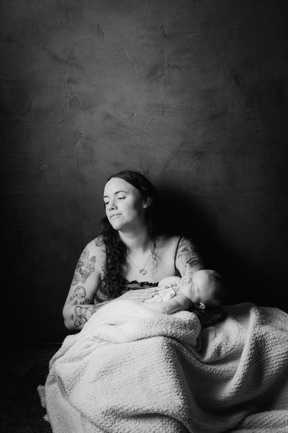 Black and white portrait of tattooed mother tenderly breastfeeding newborn baby wrapped in soft blanket.