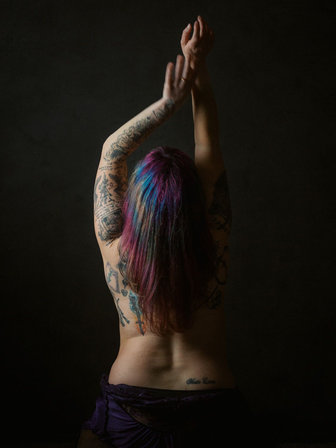 Back view of a tattooed woman with colorful pink and blue hair, arms raised, wearing a purple skirt.