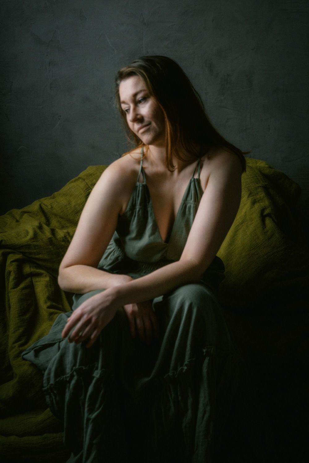 Woman in olive green dress sits relaxed on a green sofa, smiling softly in a moody studio portrait.