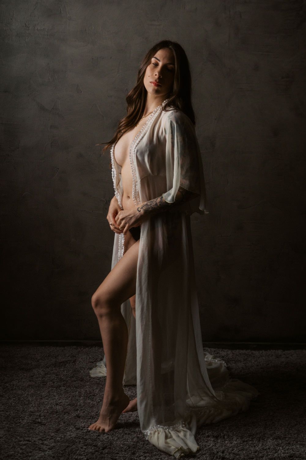 Woman in sheer white lace robe posing elegantly against dark studio backdrop in boudoir photography.
