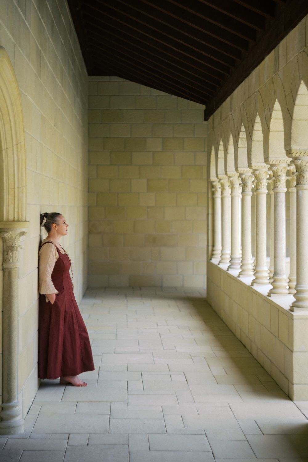 Woman in red medieval dress standing in a historic stone cloister with arched columns.
