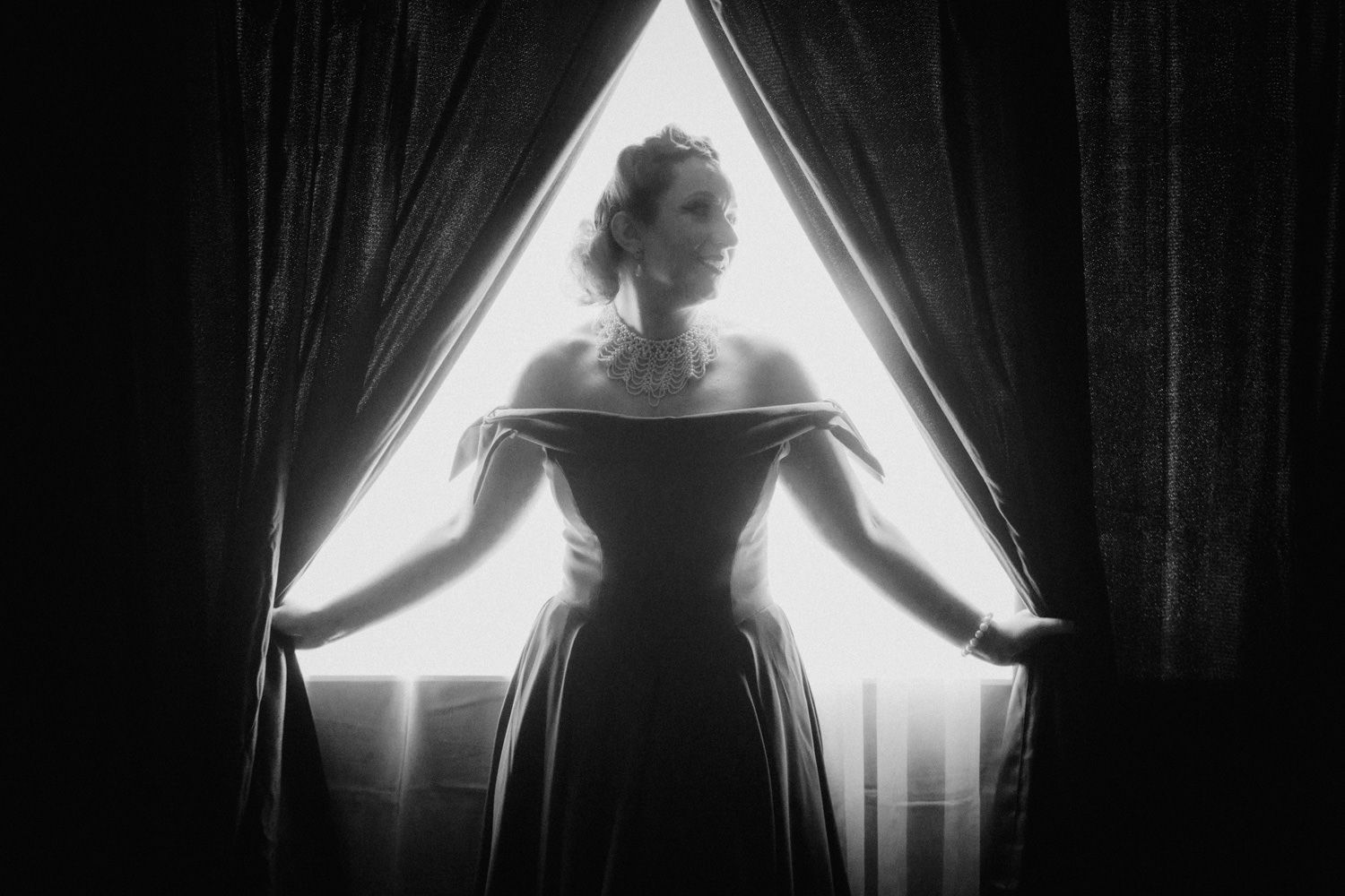 Black and white photo of a woman in an elegant off-shoulder gown standing between dramatic curtains by a bright window.