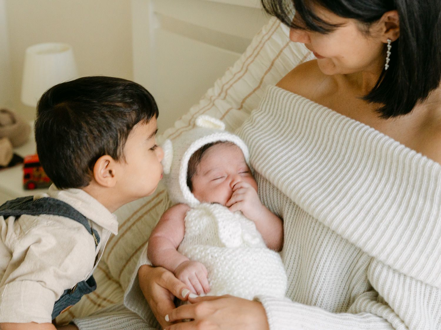 Mother in striped sweater holds sleeping newborn in bear bonnet as toddler sibling kisses baby.